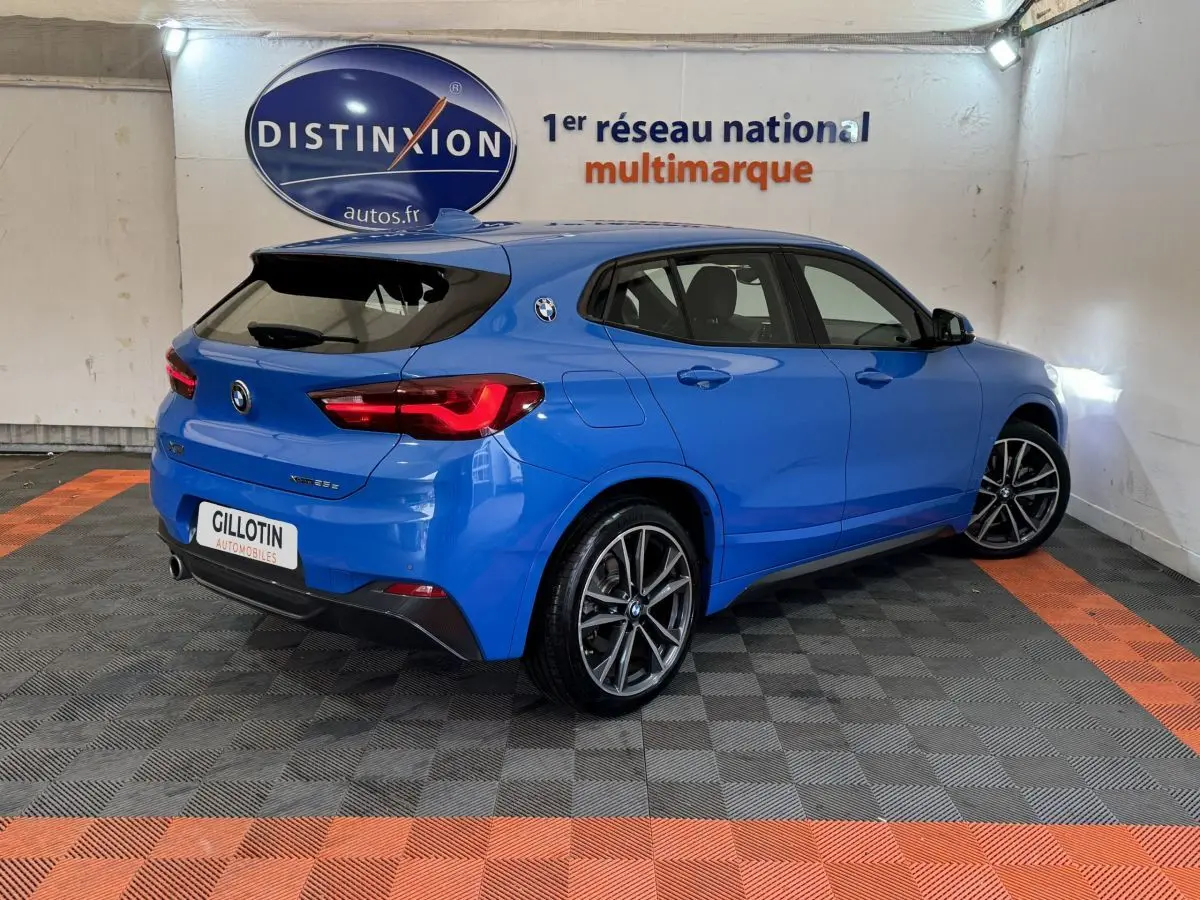 BMW X2 xDrive25e bleu en 3/4 arrière droit dans un showroom avec jantes sport et logo BMW sur l'aile arrière.