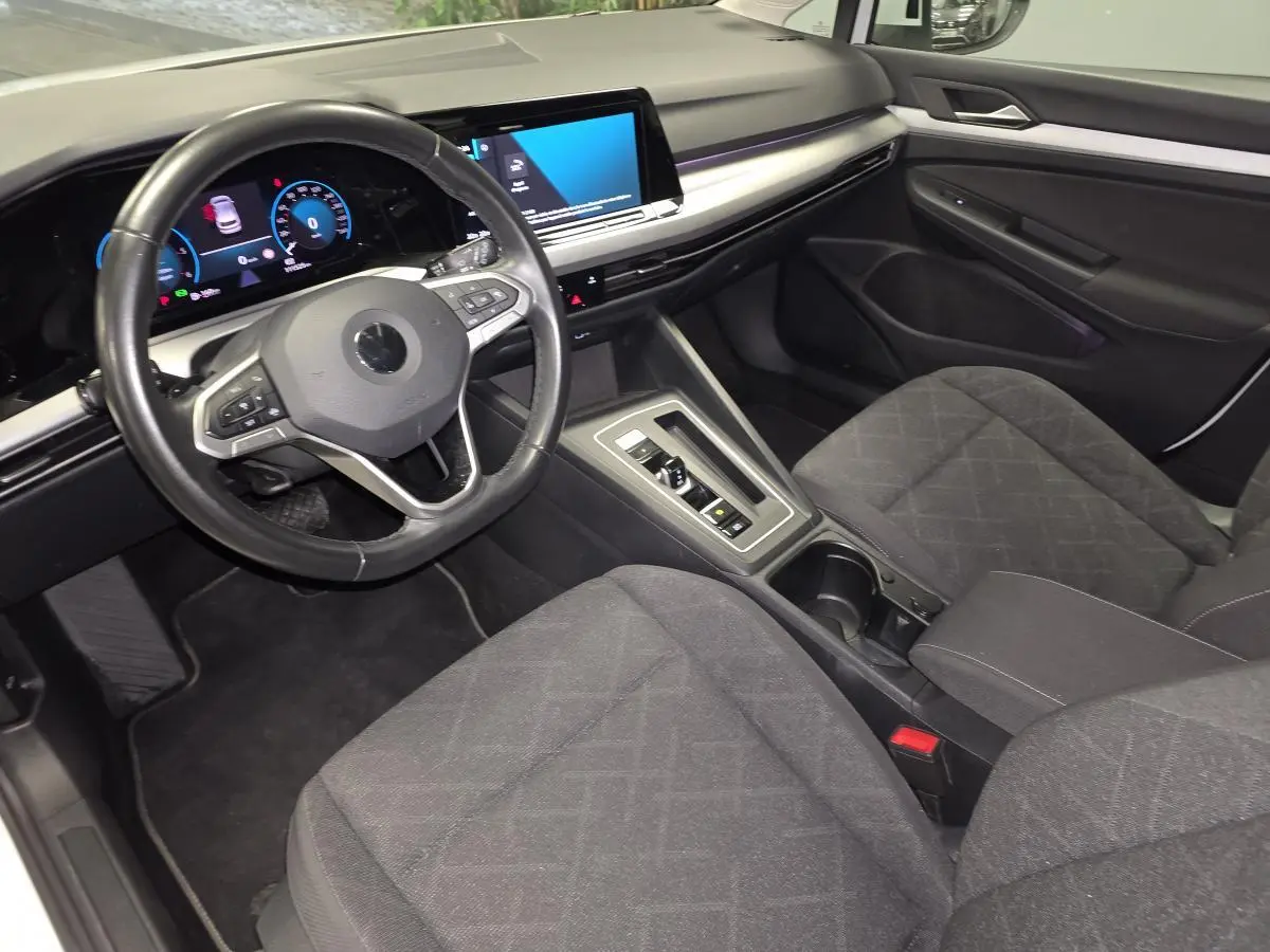 Intérieur de Volkswagen Golf VIII blanc, vue 3/4 avant côté conducteur, tableau de bord et sièges tissu gris visibles.