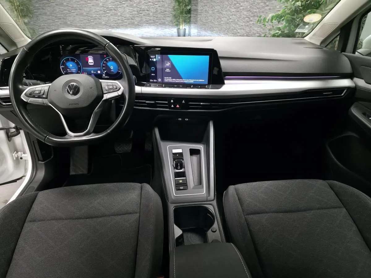 Vue intérieure avant de la Volkswagen Golf VIII blanche, tableau de bord moderne avec écran tactile et volant multifonction.