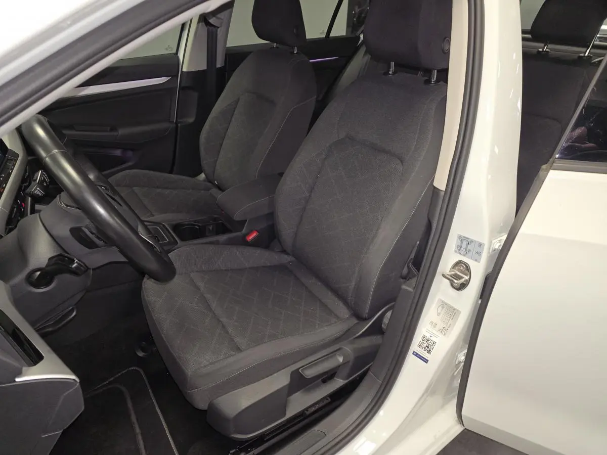 Vue intérieure côté conducteur d'une Volkswagen Golf VIII blanche, sièges avant en tissu gris foncé avec motifs.
