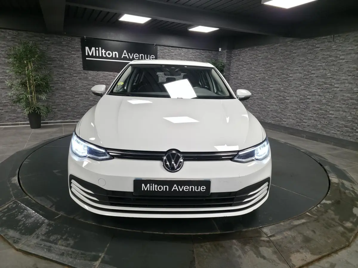 Vue avant d'une Volkswagen Golf VIII blanche avec phares LED allumés dans un showroom moderne.