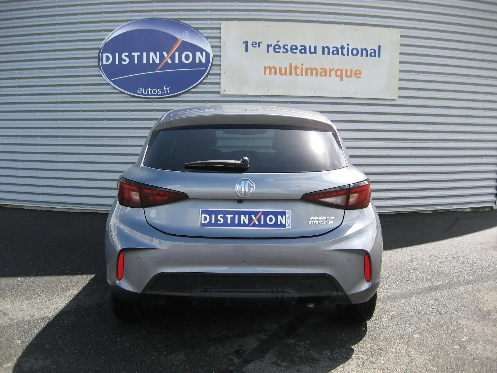 Vue arrière d'une MG3 1.5 Hybrid gris 2025 avec feux LED et logo MG bien visibles sur fond de mur métallique.