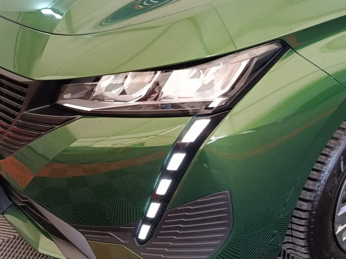 Gros plan sur l'avant droit vert métallisé d'une Peugeot 308 2023, mettant en valeur les phares LED et la calandre noire.