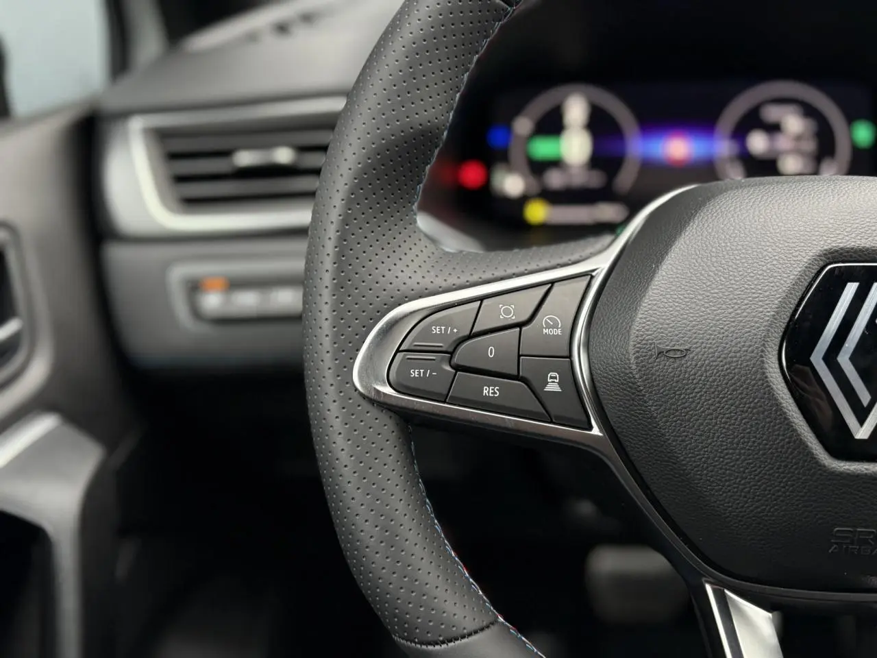 Gros plan sur le volant perforé avec commandes intégrées et logo Alpine du Renault Captur Blanc Nacré, tableau de bord numérique flou en arrière-plan.