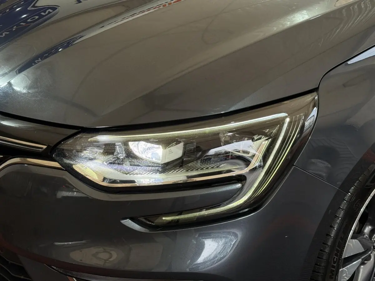 Gros plan sur le phare avant droit d’une Renault Mégane Intens gris foncé, avec détails LED et partie de la calandre visible.