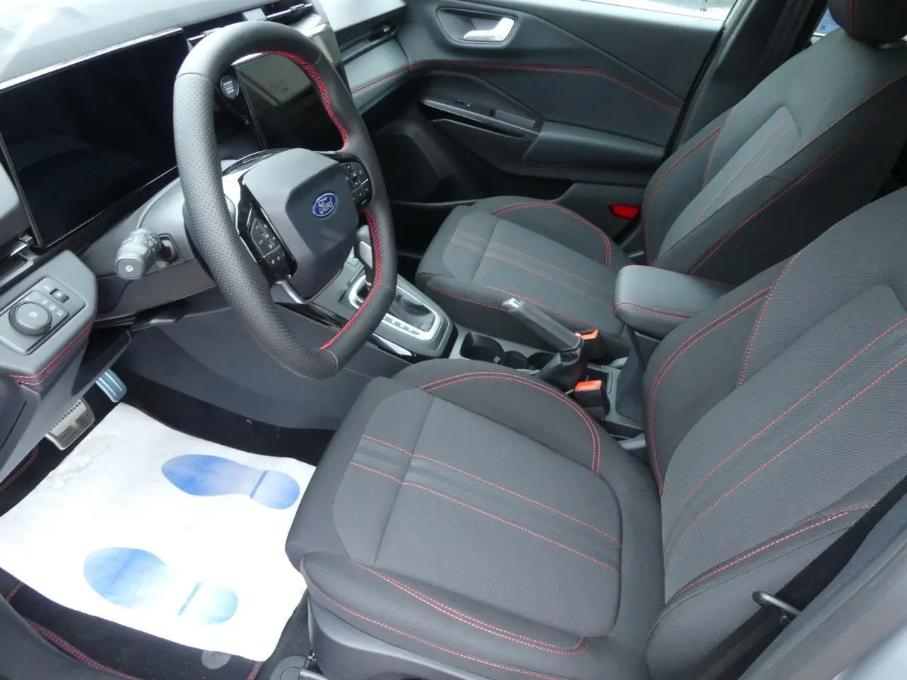 Vue intérieure côté conducteur du Ford Puma 2025, sièges tissu gris avec surpiqûres rouges et volant sport à coutures rouges.