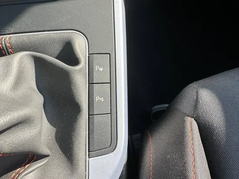 Gros plan intérieur sur la console centrale du SEAT Arona 2025, montrant le soufflet de levier de vitesses noir avec surpiqûres rouges et boutons d’aide au stationnement.