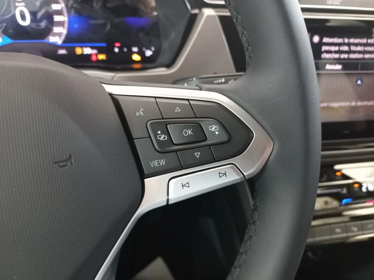Gros plan sur le côté gauche du volant noir du Volkswagen Touran 2024 avec commandes multifonctions et affichage numérique en arrière-plan.