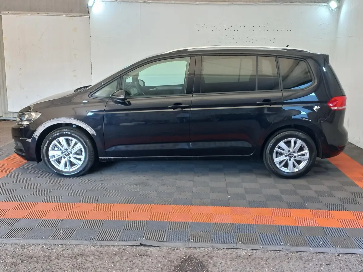 Vue de profil côté gauche d’un Volkswagen Touran noir 7 places, sur sol à damier gris et orange en intérieur.