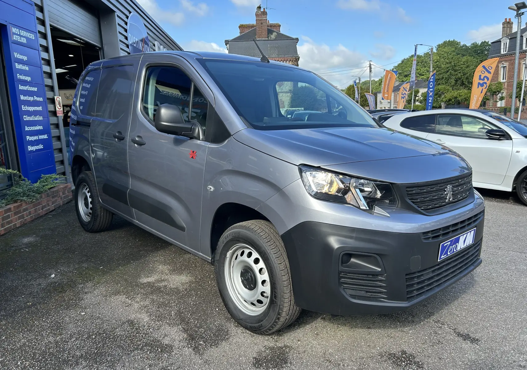 Vue 3/4 avant d'un Peugeot Partner Dangel 4x4 gris Artense avec protections noires et jantes acier, stationné devant un garage.