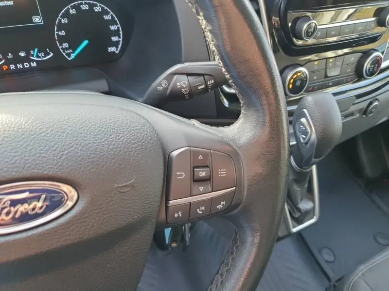 Vue rapprochée du volant et du tableau de bord du Ford Transit Custom blanc glacier, avec commandes multifonctions et levier de vitesses automatique.