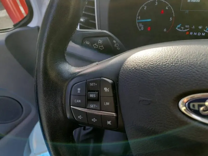 Gros plan sur le volant noir du Ford Transit Custom blanc, montrant les commandes de régulateur et l'instrumentation partielle.