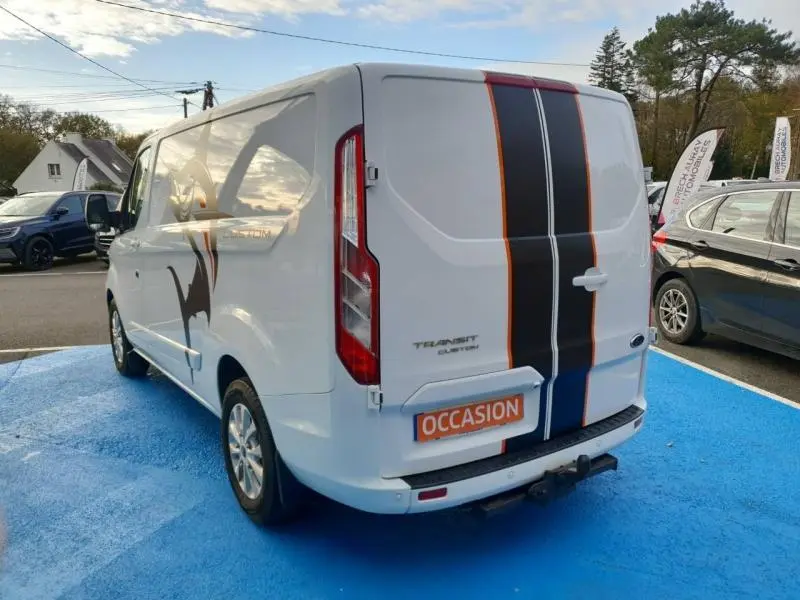 Vue 3/4 arrière droite du Ford Transit Custom blanc Glacier avec bandes noires et orange et crochet d'attelage visible.