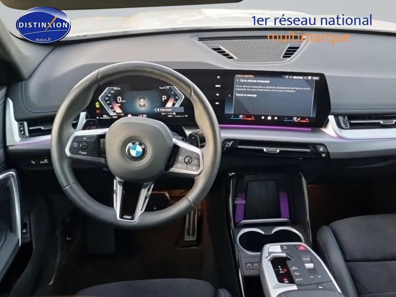 Vue intérieure frontale du tableau de bord et volant cuir noir du BMW X1 blanc minéral 2025 avec écran tactile et éclairage d'ambiance violet.