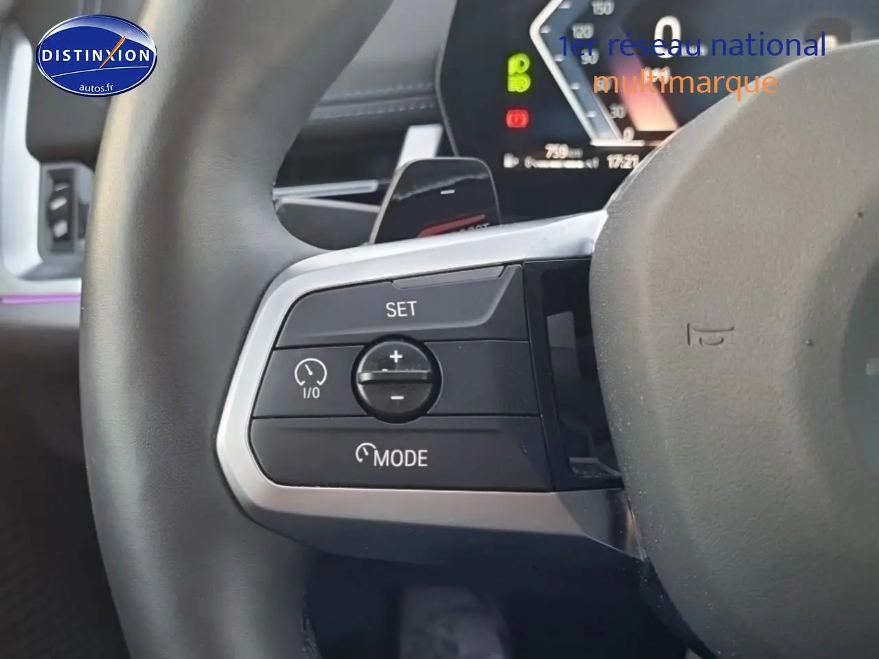 Gros plan sur les commandes de régulateur de vitesse au volant cuir du BMW X1 blanc mineral, tableau de bord numérique en arrière-plan.