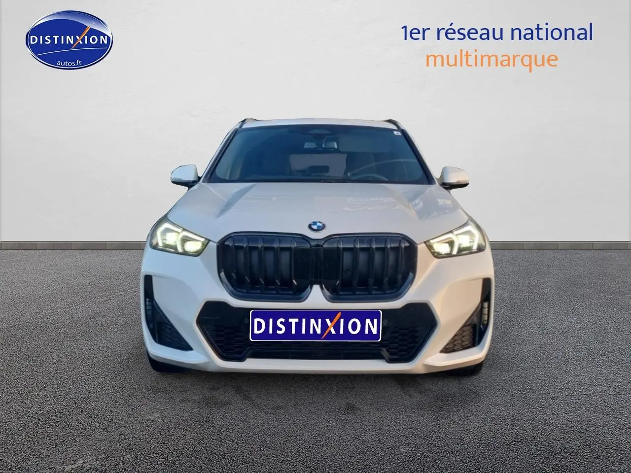 Vue frontale d'un BMW X1 blanc minéral avec calandre noire et phares LED allumés sur fond neutre.