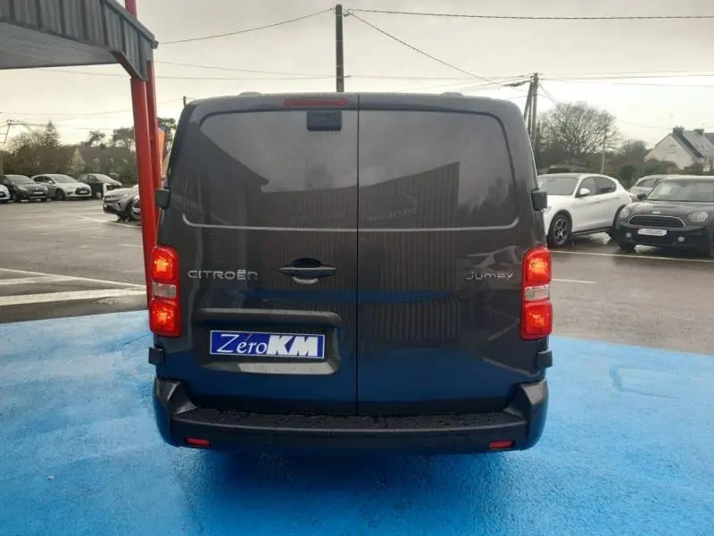 Vue arrière d'un Citroën Jumpy gris titane métal avec portes battantes et feux arrière allumés sur parking.