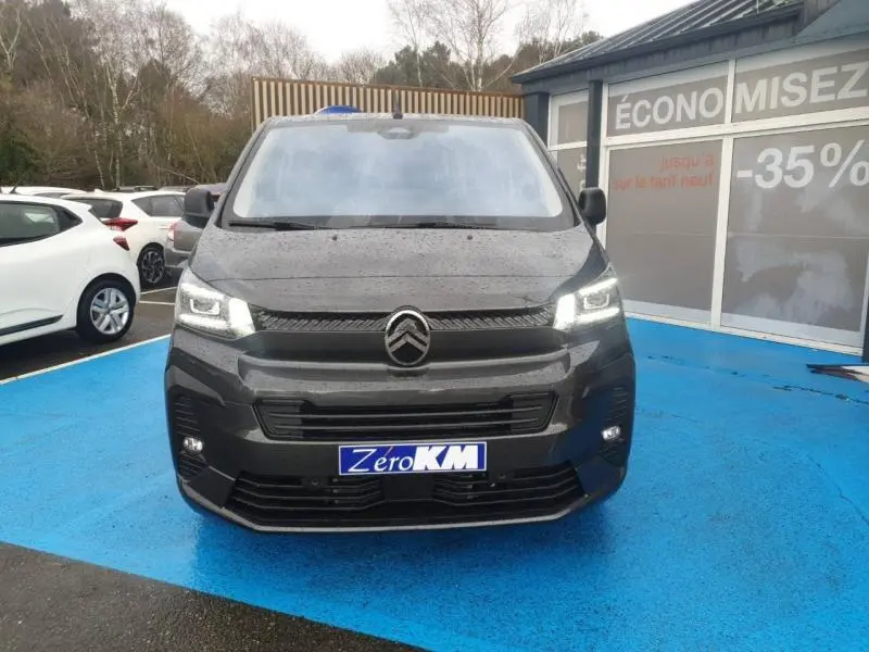Vue avant d'un Citroën Jumpy gris titane métal 2025 avec projecteurs LED allumés sur parking bleu.
