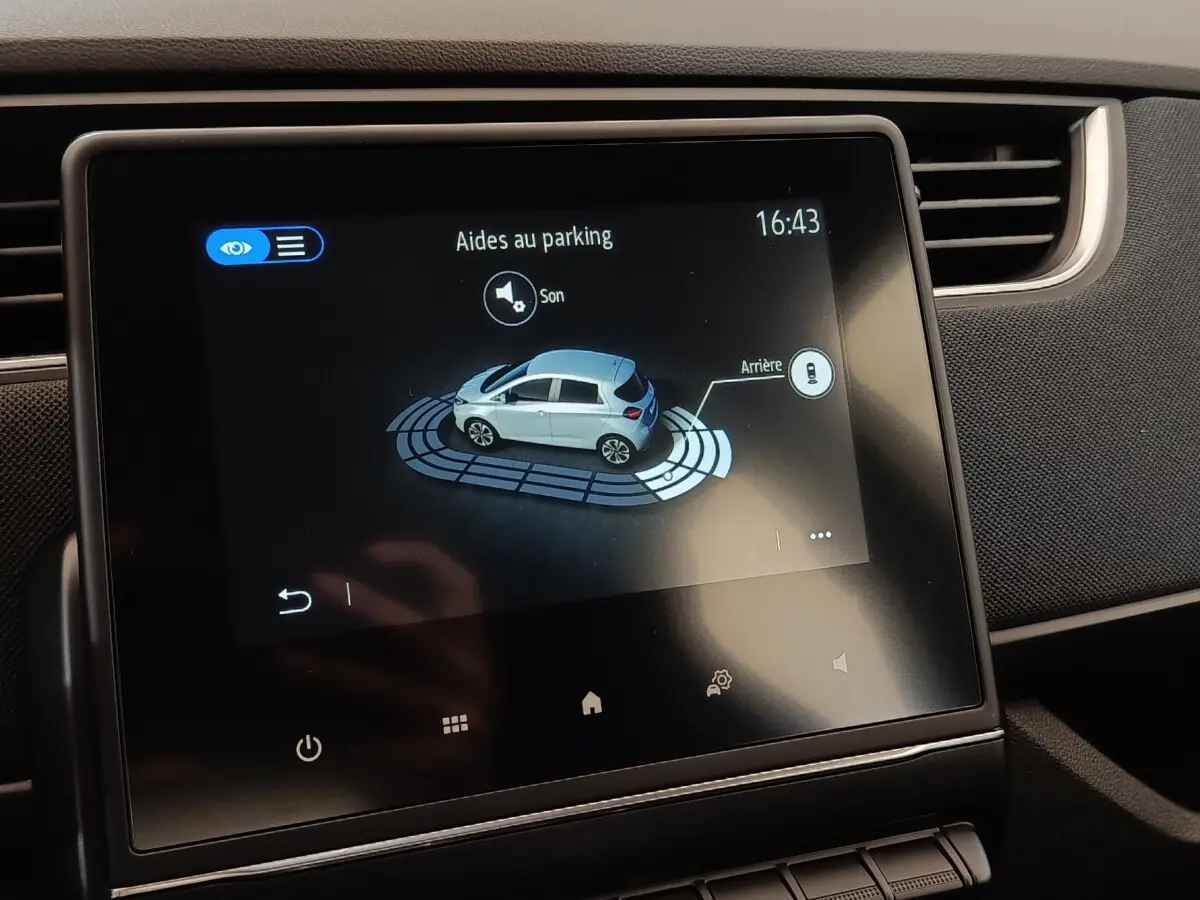 Écran tactile central affichant l’aide au parking d’une Renault ZOE blanche vue de profil côté gauche.