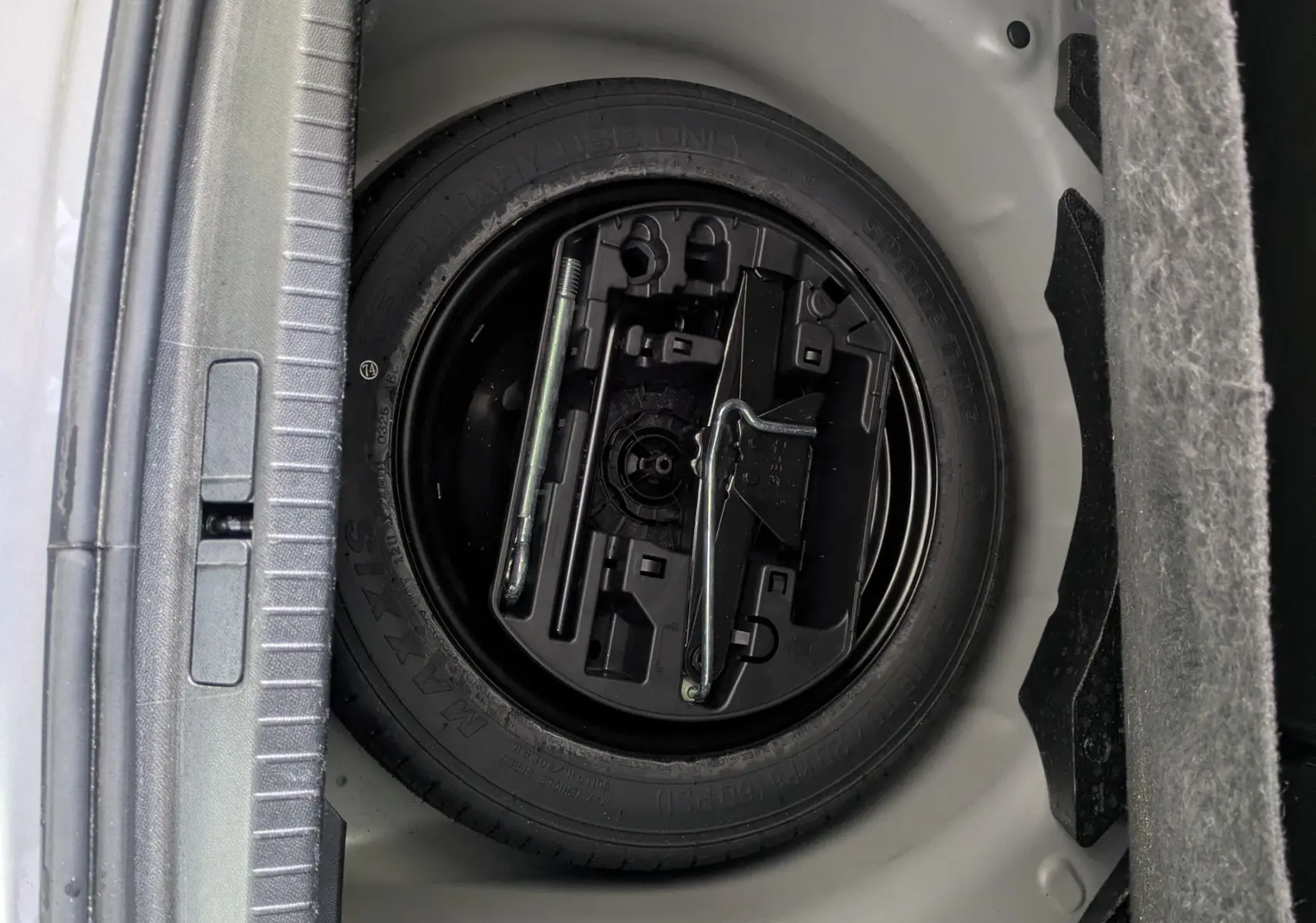 Vue rapprochée du coffre d'une Peugeot 208 Hybrid 145 gris Artense, montrant la roue de secours et les outils de bord.