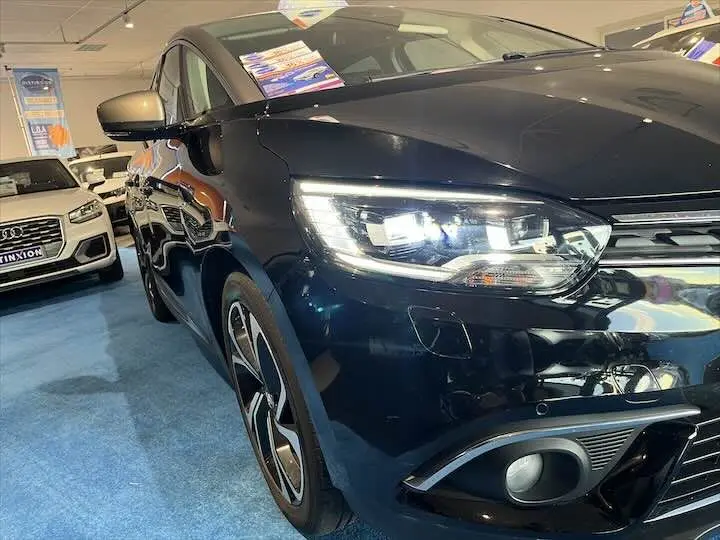 Vue 3/4 avant gauche d'un Renault Scénic IV noir étoile métal avec toit gris, phares LED allumés en showroom.