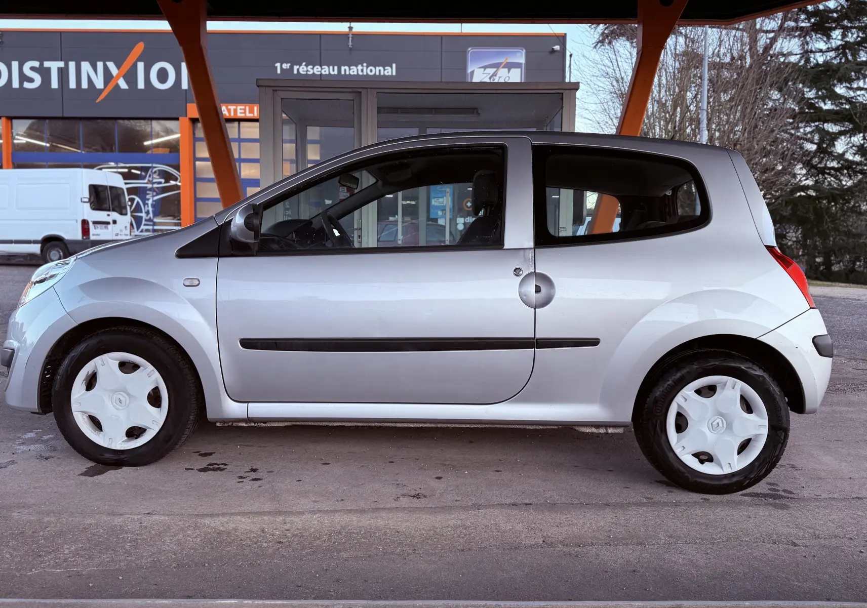 Profil côté gauche d'une Renault Twingo II grise 3 portes, avec jantes blanches et poignées noires visibles.