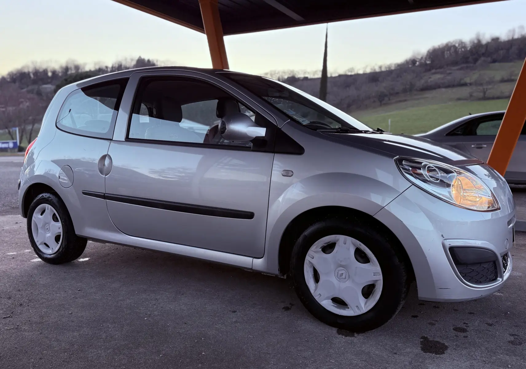 Renault Twingo II gris vue de profil côté gauche sous abri, phare avant droit allumé, jantes avec enjoliveurs blancs.