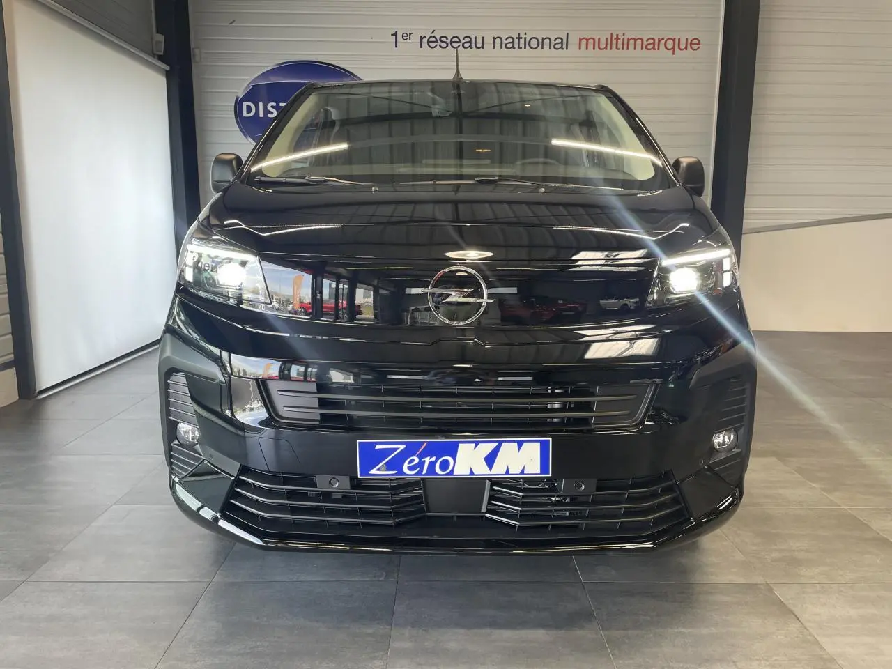 Vue frontale d'un Opel Vivaro noir carbone avec phares allumés et plaque Zéro KM visible en intérieur showroom.