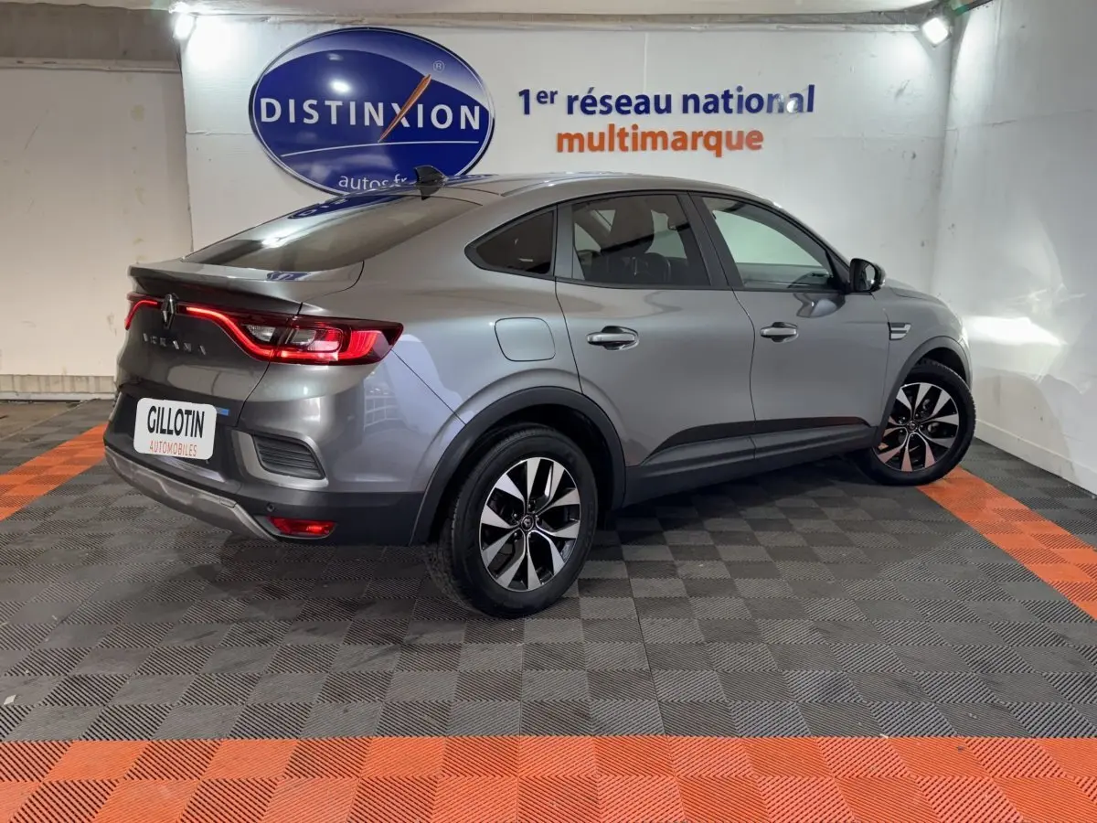 Vue 3/4 arrière droite d'une Renault Arkana Business E-TECH 145 gris foncé avec feux arrière LED allumés en intérieur showroom.