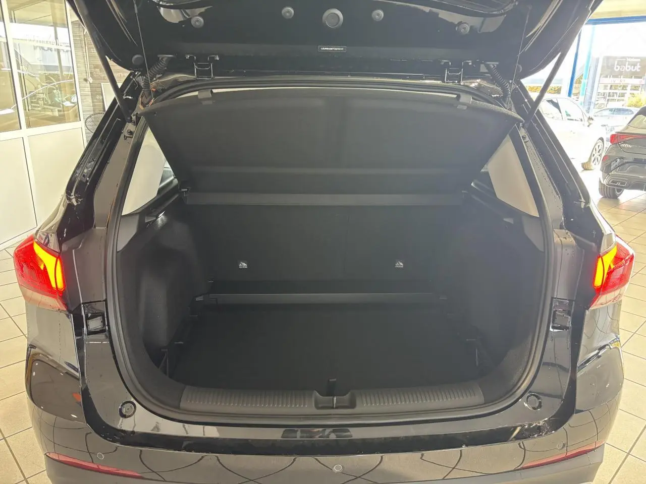 Coffre ouvert en vue arrière d’un MG Motor ZS 1.5 Hybrid+ noir, avec feux arrière allumés en intérieur showroom.