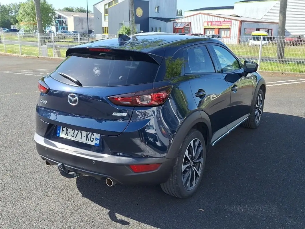 Vue 3/4 arrière droite d'un Mazda CX-3 bleu avec jantes aluminium et attelage visible sur parking.