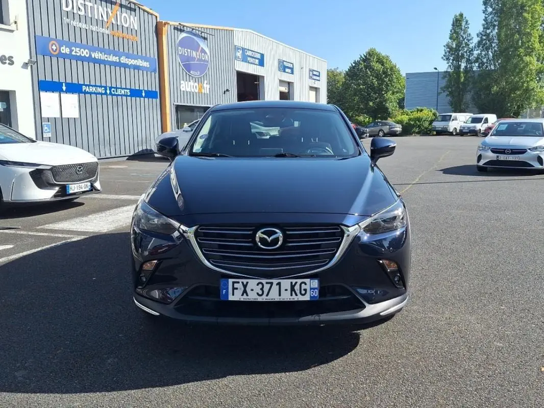 Vue avant d'un Mazda CX-3 bleu foncé garé devant un garage, avec calandre chromée et phares allumés.