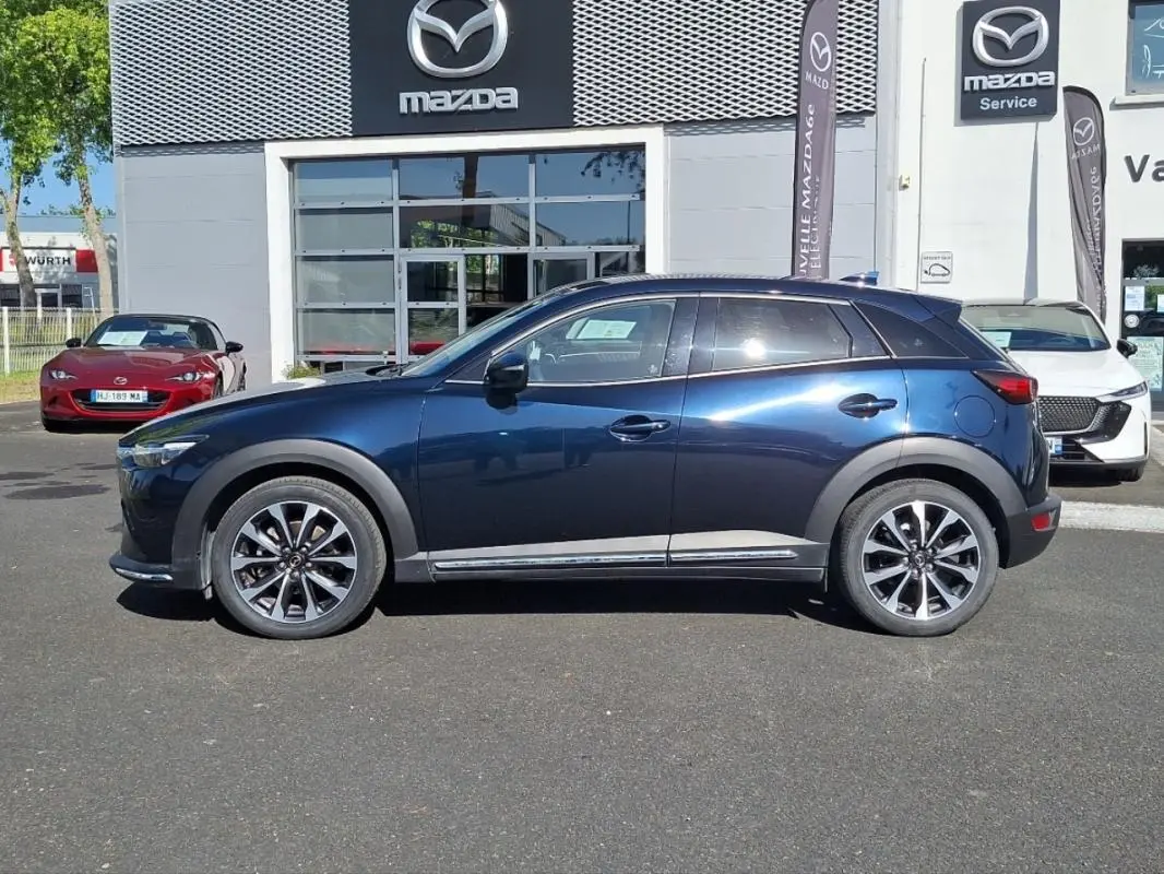 Vue de profil côté gauche d'un Mazda CX-3 bleu 2021 stationné devant un concessionnaire Mazda.
