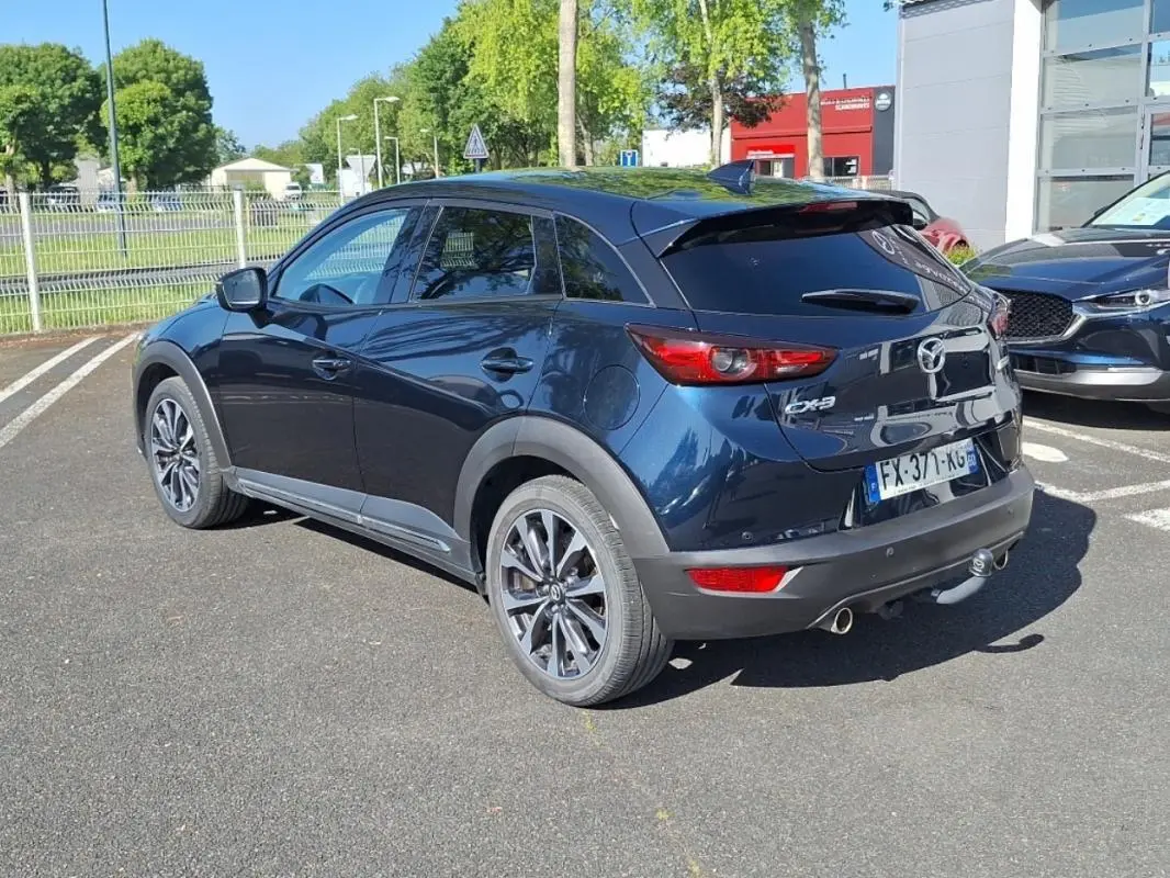 Vue 3/4 arrière droite d'un Mazda CX-3 bleu 2021 avec jantes aluminium et attelage visible en extérieur.