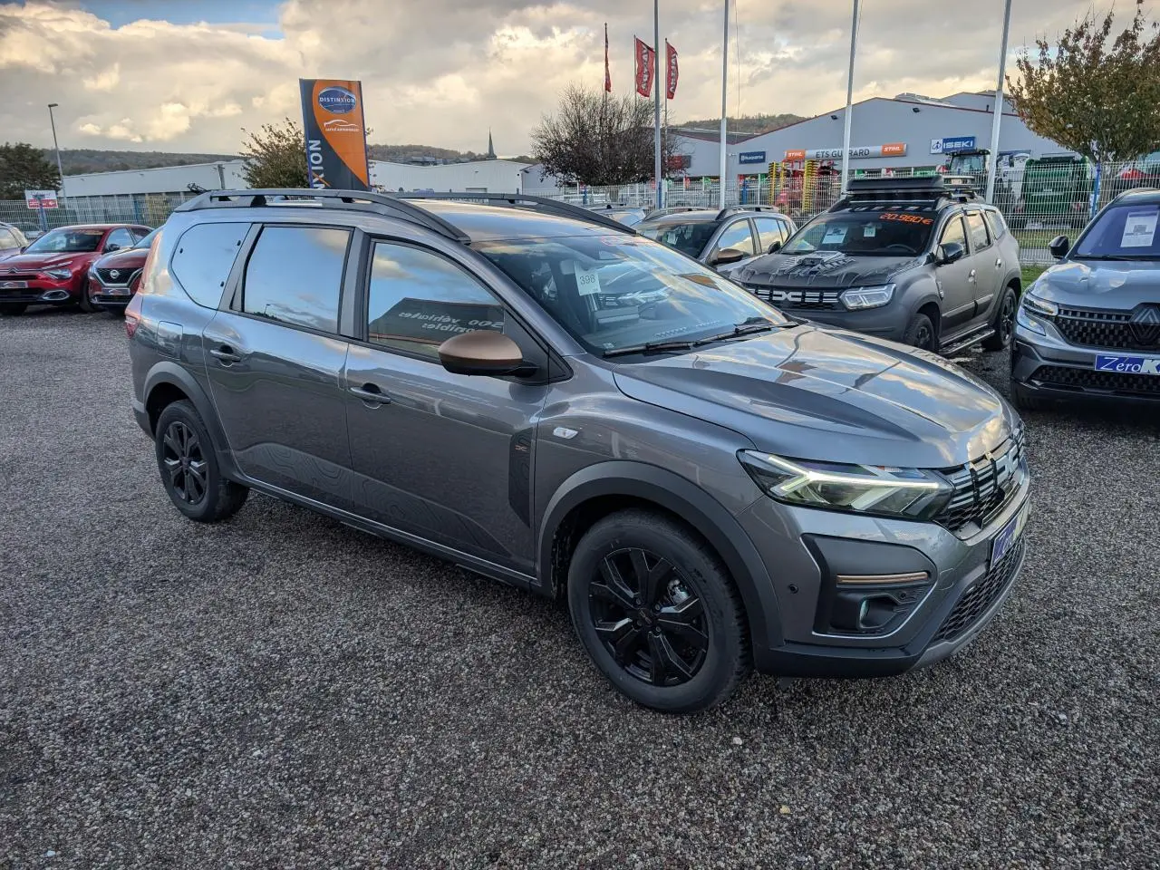 Dacia Jogger gris métallisé en 3/4 avant droit avec jantes noires et barres de toit sur parking.