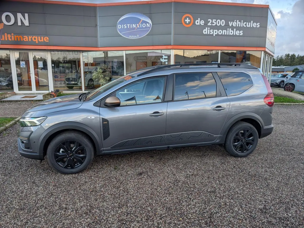 Vue de profil côté gauche d’un Dacia Jogger gris métallisé avec jantes noires et rétroviseurs brun cuivré, stationné devant un concessionnaire.