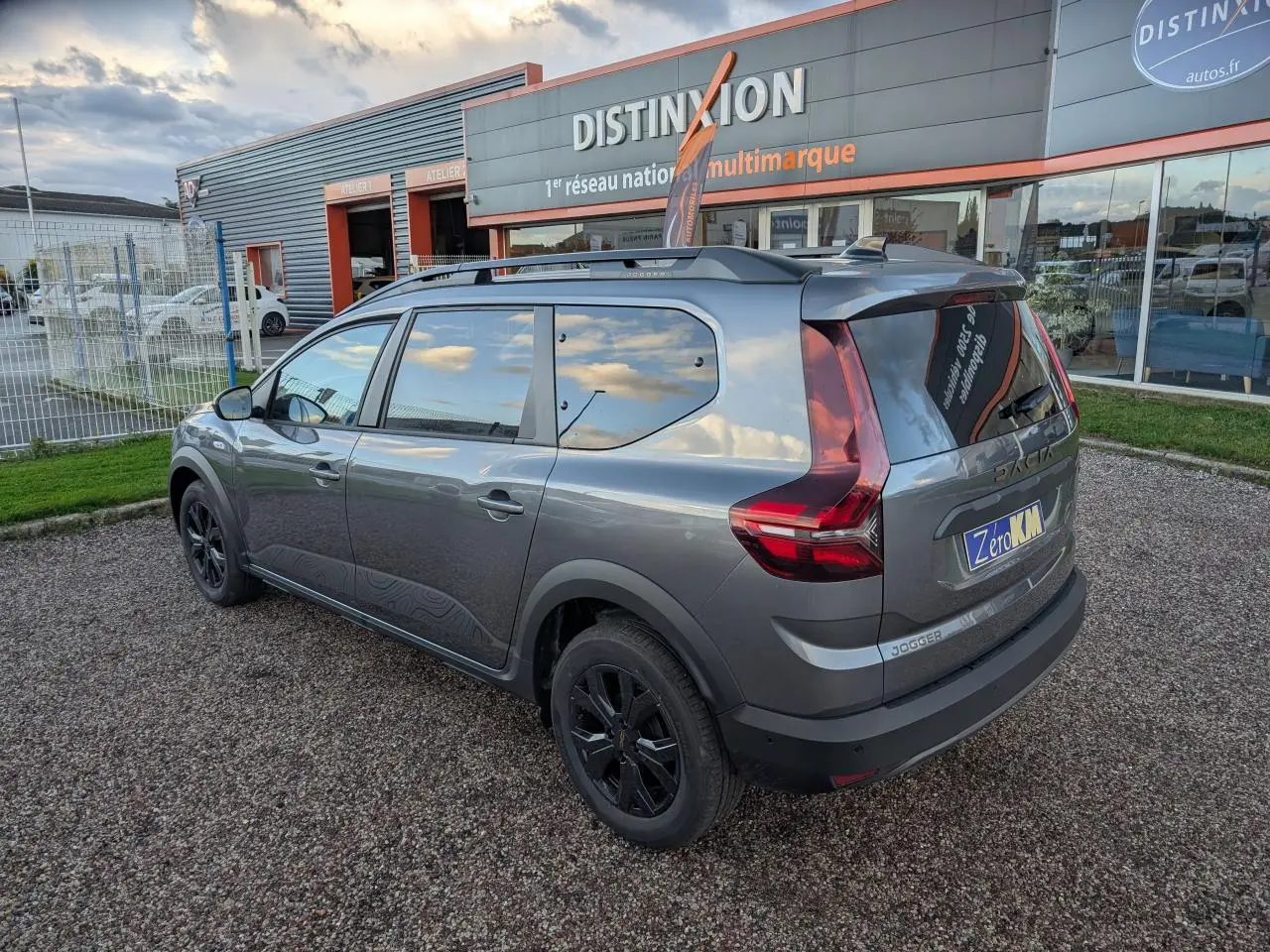 Vue 3/4 arrière droite d'un Dacia Jogger gris métallisé avec jantes noires et barres de toit, stationné devant un garage.