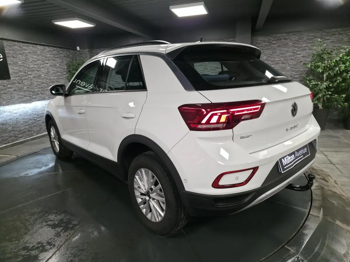 Vue 3/4 arrière droite du Volkswagen T-Roc blanc avec attelage amovible et feux arrière LED allumés en intérieur showroom.