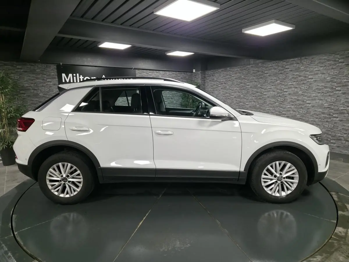 Volkswagen T-Roc blanc vu de profil côté gauche, avec jantes alliage et toit ouvrant en intérieur showroom.