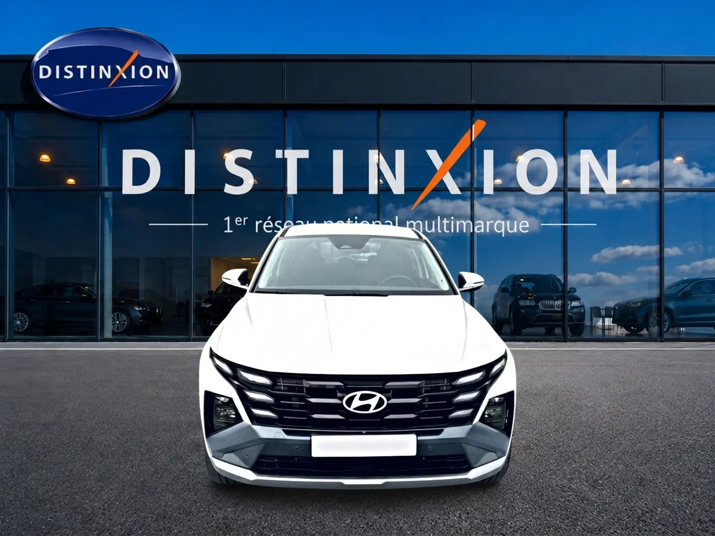Vue avant d'un Hyundai Tucson blanc 2026 avec calandre noire distinctive, garé devant une vitrine de concession multimarque.