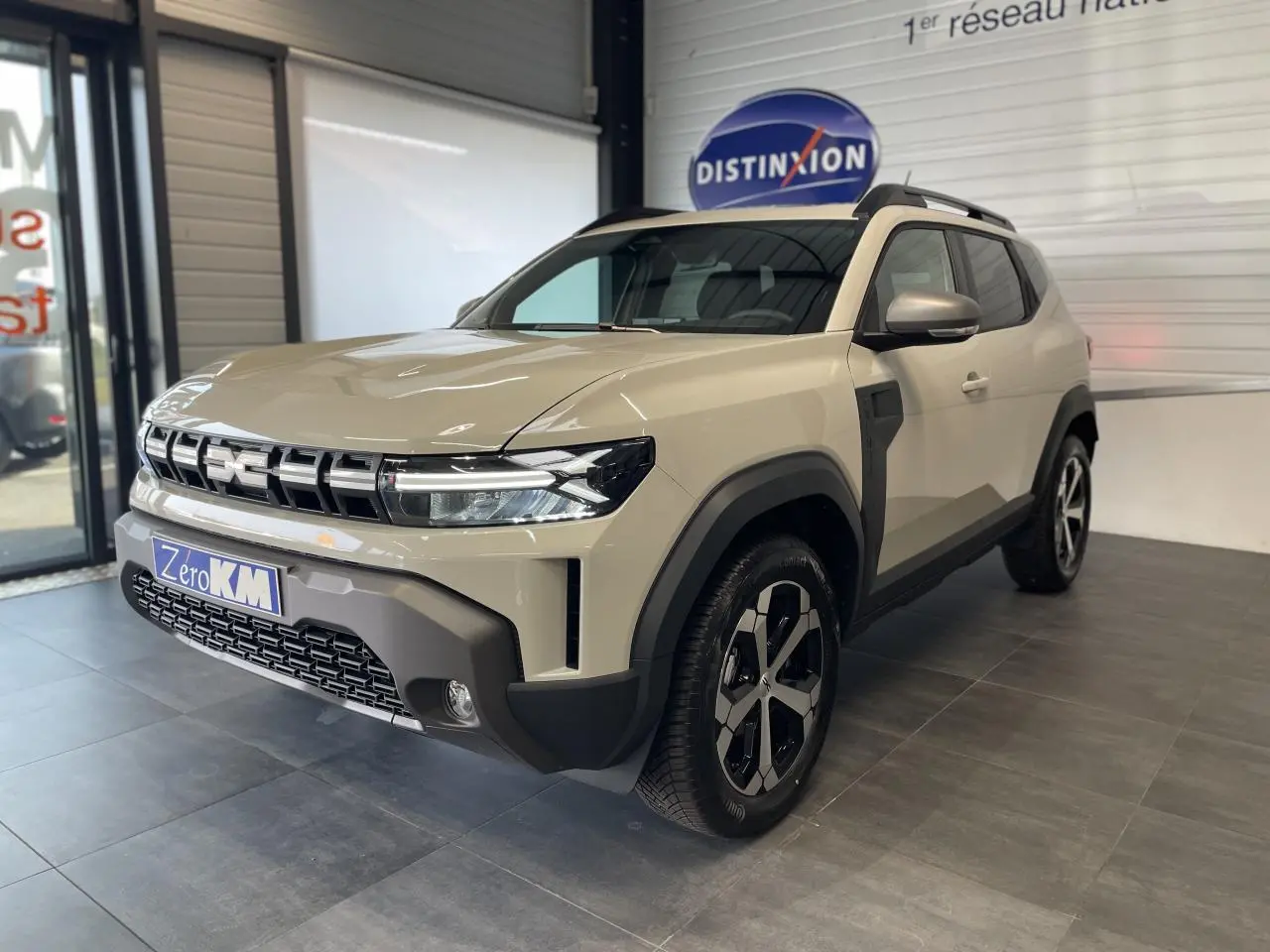 Vue 3/4 avant d'un Dacia Duster beige Sandstone 2025 avec jantes alliage 18 pouces et barres de toit noires.