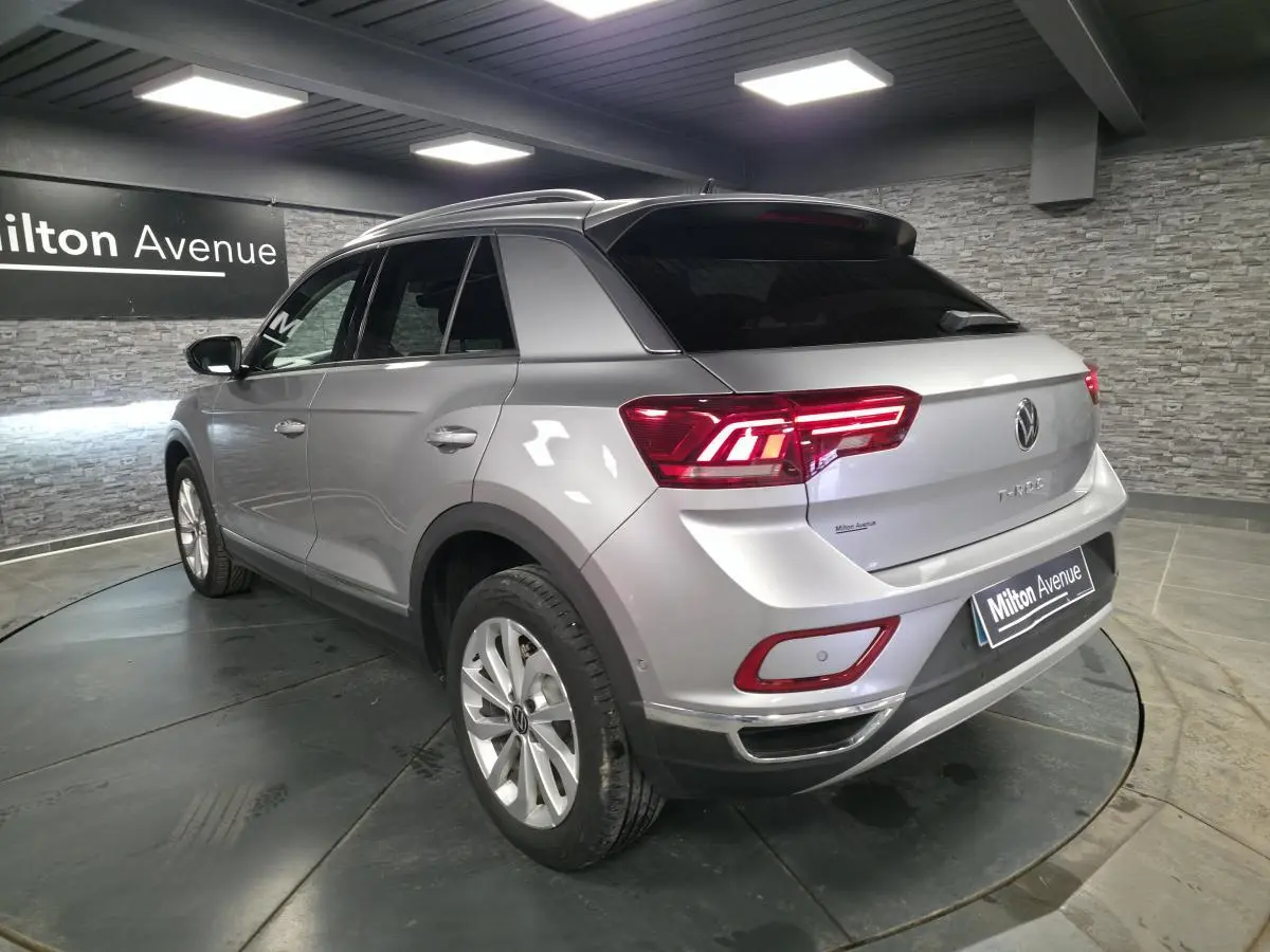 Vue 3/4 arrière droite d'un Volkswagen T-Roc gris métallisé avec feux arrière LED allumés en intérieur showroom.