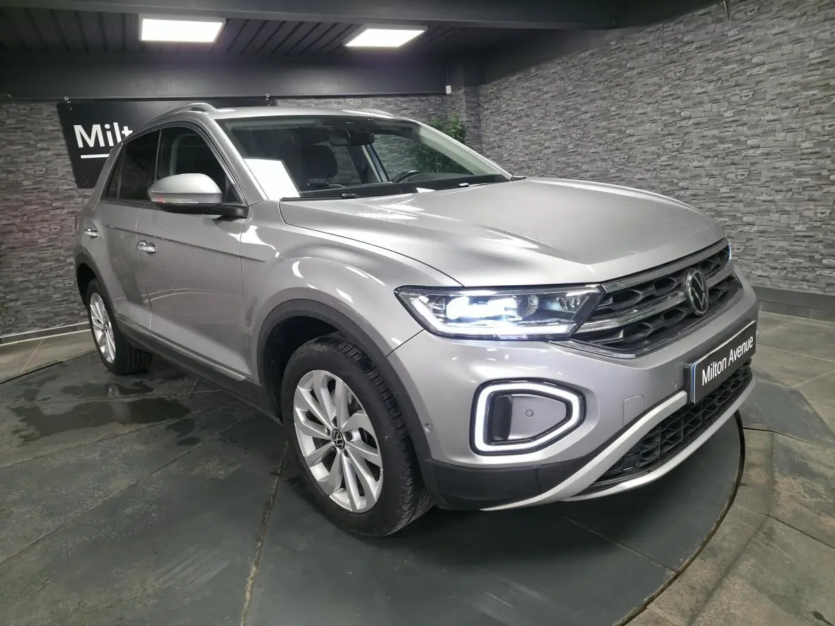 Volkswagen T-Roc gris métallisé vue 3/4 avant droit avec feux LED allumés en intérieur showroom.