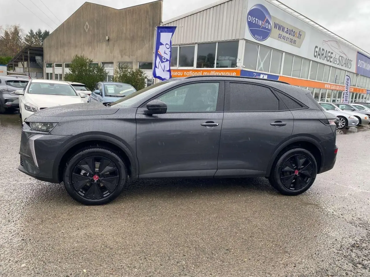 Vue de profil côté gauche du DS7 Crossback gris foncé 2024 avec jantes noires et vitres teintées, sur parking extérieur humide.