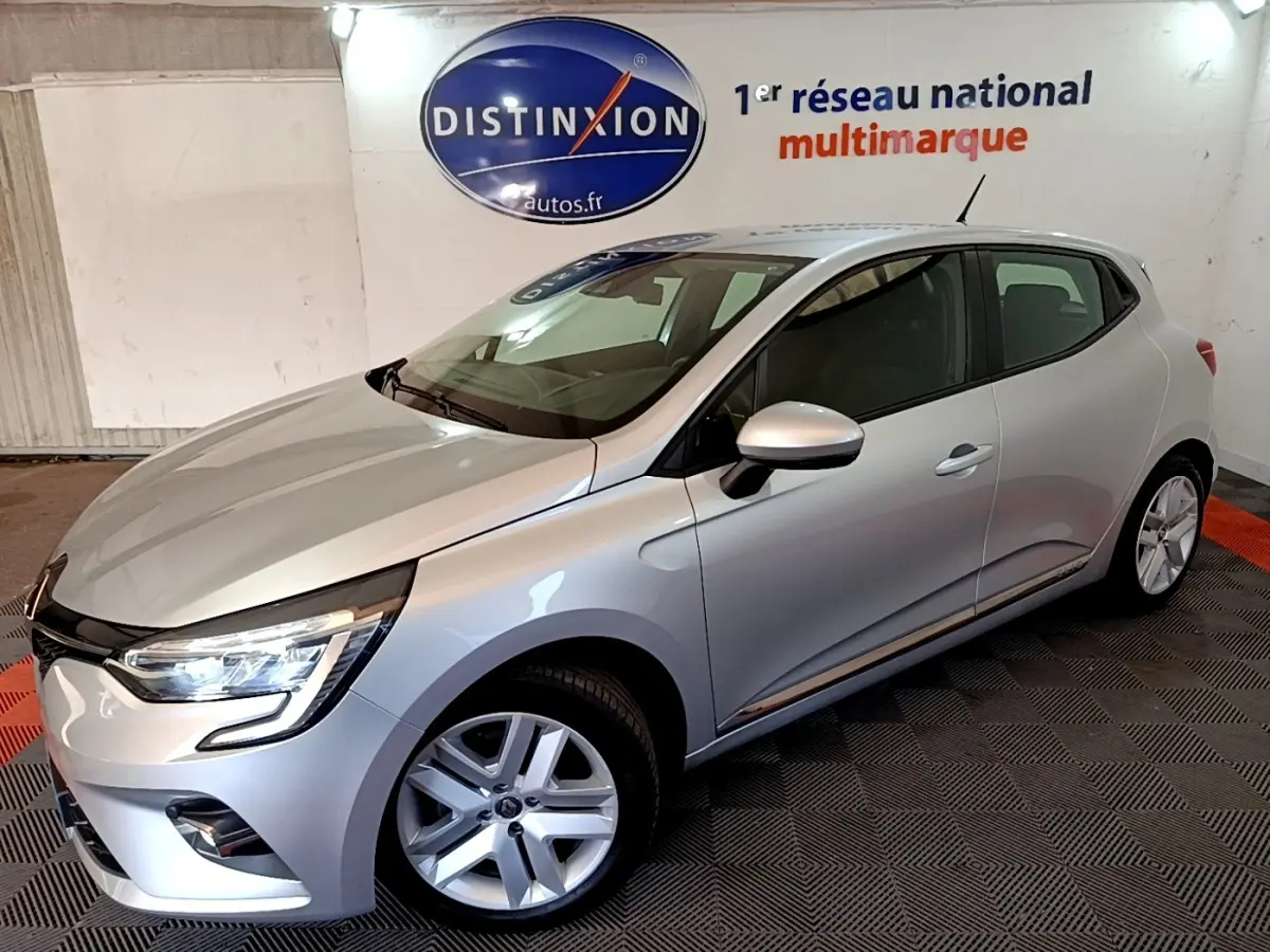 Vue 3/4 avant droite d'une Renault Clio gris clair avec feux LED allumés dans un showroom intérieur.