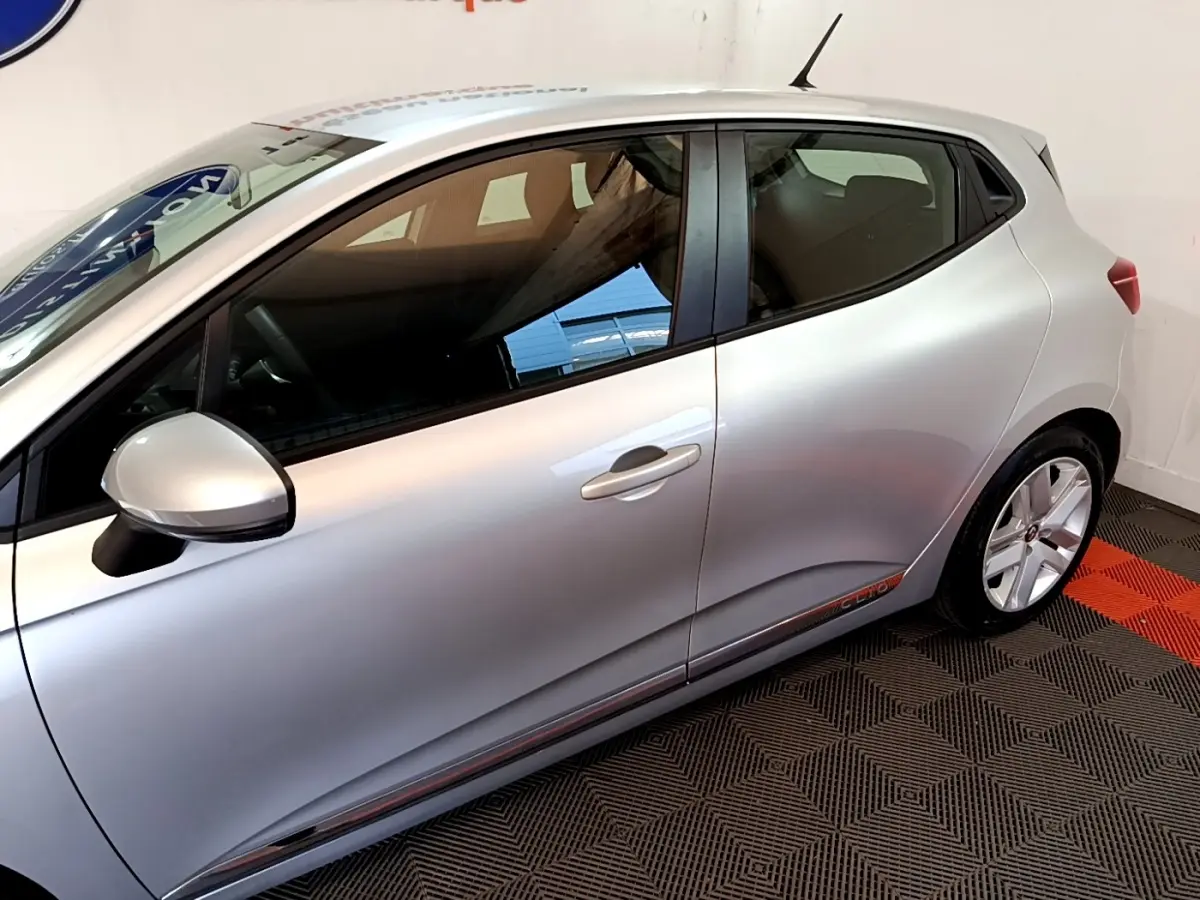 Profil droit de la Renault Clio gris clair avec poignées intégrées et jantes alliage visibles en intérieur showroom.