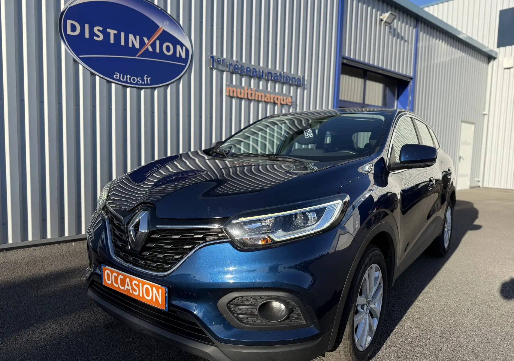 Renault Kadjar bleu 2019 en 3/4 avant droit, avec calandre noire et feux LED, stationné devant un bâtiment gris.