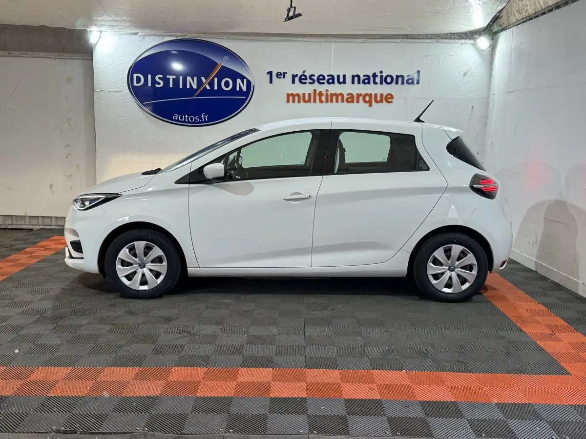 Profil droit d'une Renault ZOE blanche 2020 électrique, avec jantes simples et feux arrière allumés en intérieur showroom.