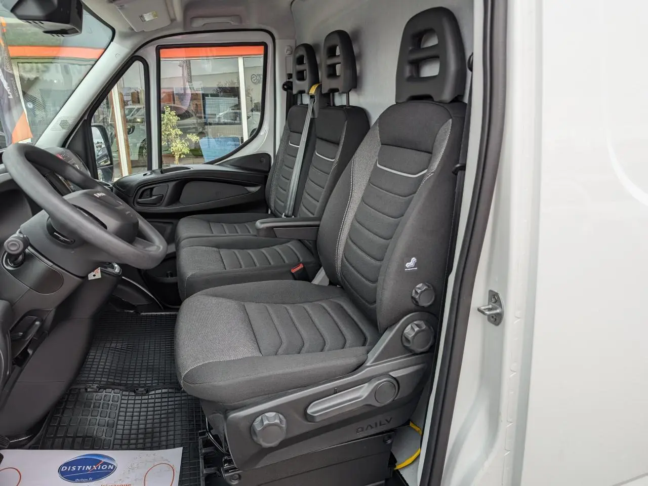 Vue intérieure côté conducteur du fourgon Iveco Daily blanc 2025, montrant les sièges en tissu gris et le volant.