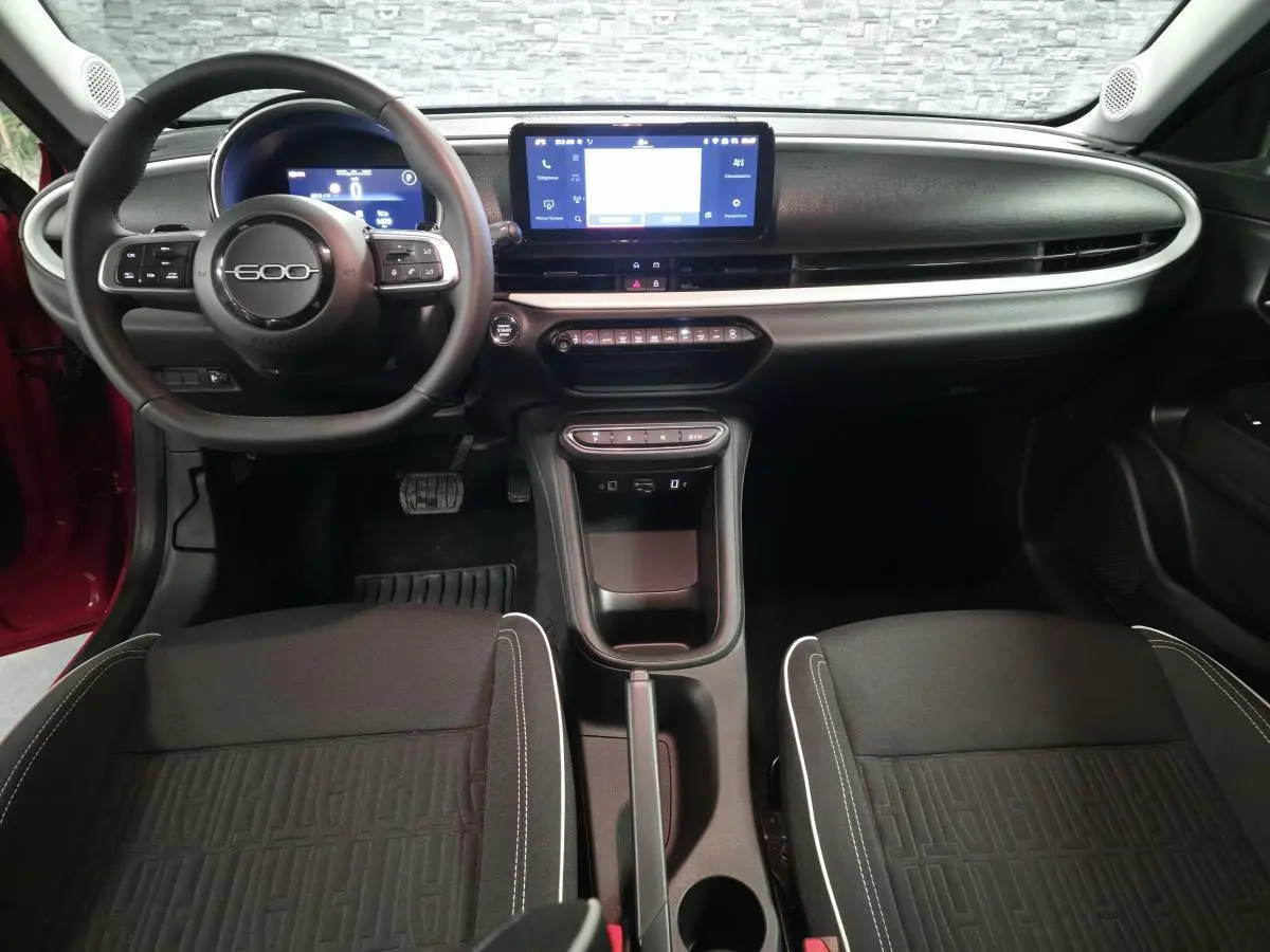 Vue intérieure avant de la FIAT 600 rouge 2024, tableau de bord noir mat avec écran tactile central et volant multifonction.